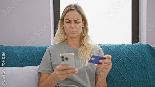 A perplexed young woman examines her credit card while using a smartphone in her cozy living room.