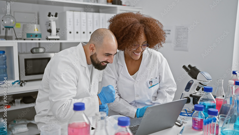 Two amiable scientists happily speaking and discovering breakthroughs together on laptop in busy laboratory amidst test tubes and microscopes Two amiable scientists happily speaking and discovering breakthroughs together on laptop in busy laboratory amidst test tubes and microscopes