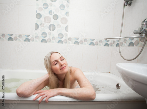 A beautiful smiling sexy girl with blond hair sits in a bath and washes, in the bathroom. An image about cleanliness and about taking care of yourself and your body.
