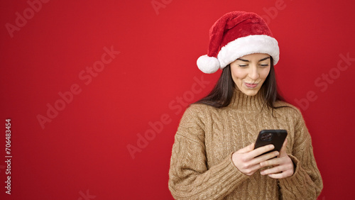 Young beautiful hispanic woman wearing christmas hat using smartphone over isolated red background