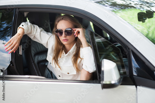 A fashionable beautiful slender blonde girl in sunglasses, light shorts and a vest sits in a black and white car. Image for fashion bloggers.
