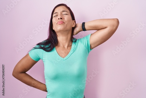 Young hispanic woman standing over pink background suffering of neck ache injury, touching neck with hand, muscular pain