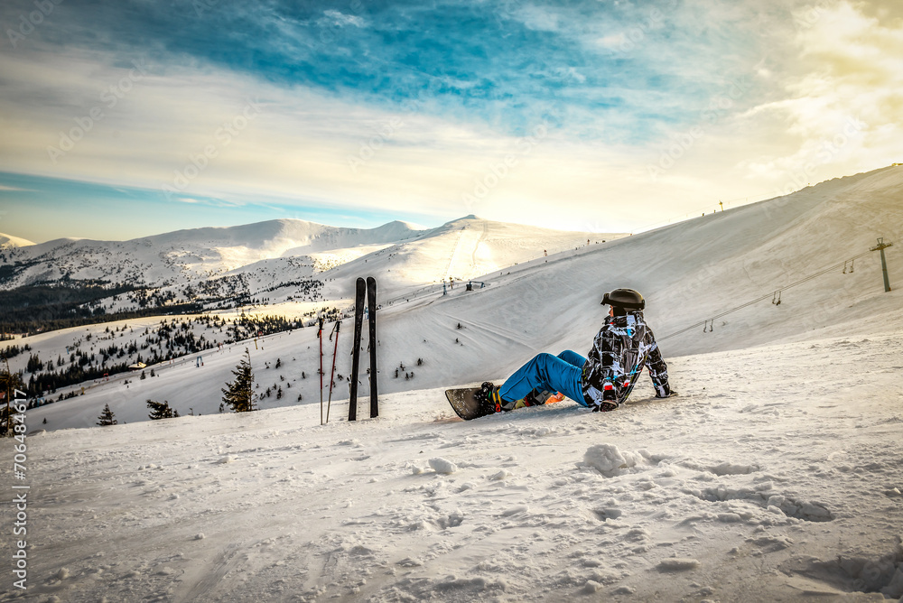 Snowboarder sits high in the mountains on the edge of the slope and looks into the distance at sunset. Winter vacation theme