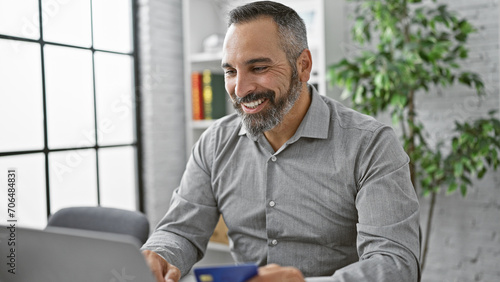Smiling mature hispanic man with gray beard working on laptop in modern office, holding credit card. photo