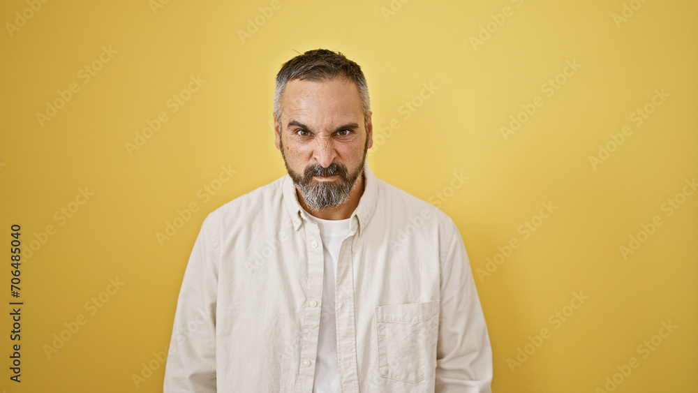 Fototapeta premium A mature hispanic man with grey beard stands confidently against a yellow wall, portraying serious expression.