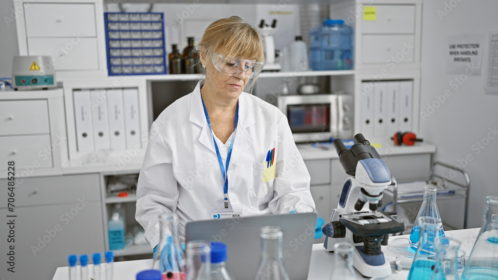 Beautiful middle age blonde woman scientist, serious and professional, analyzing results on laptop at a busy lab Beautiful middle age blonde woman scientist, serious and professional, analyzing results on laptop at a busy lab