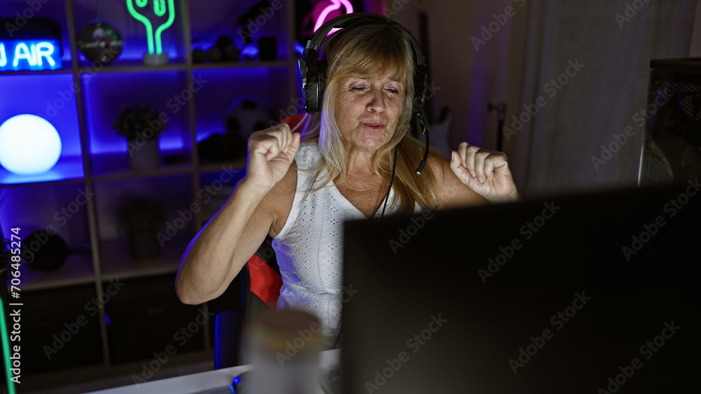 Cheerful middle age blonde woman streamer confidently smiling, playfully winning video game on stream; feel-good moment in the dark gaming room, embodying virtual triumph! Cheerful middle age blonde woman streamer confidently smiling, playfully winning video game on stream; feel-good moment in the dark gaming room, embodying virtual triumph!