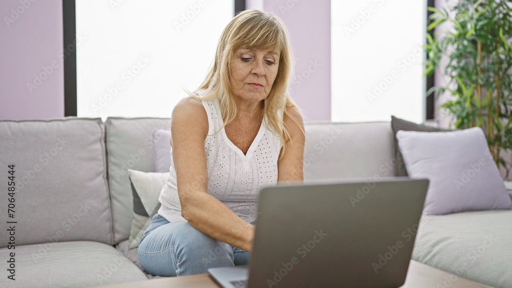 In her cozy home, a serious and focused middle age blonde woman is sitting on the sofa, totally absorbed, using her laptop in the living room's welcoming interior. In her cozy home, a serious and focused middle age blonde woman is sitting on the sofa, totally absorbed, using her laptop in the living room's welcoming interior.
