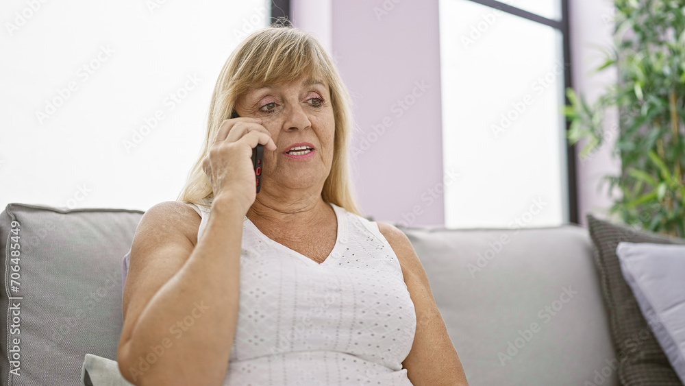 Serious middle-aged blonde woman engrossed in a serious phone talk on her smartphone, sitting upset on her living room sofa, dealing with a problem indoors at her comfortable apartment. Serious middle-aged blonde woman engrossed in a serious phone talk on her smartphone, sitting upset on her living room sofa, dealing with a problem indoors at her comfortable apartment.