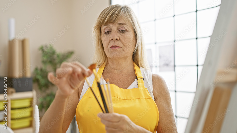 Beautiful middle-aged blonde woman artist, absorbed in choosing the right paintbrush, immerses herself in creativity within her art studio