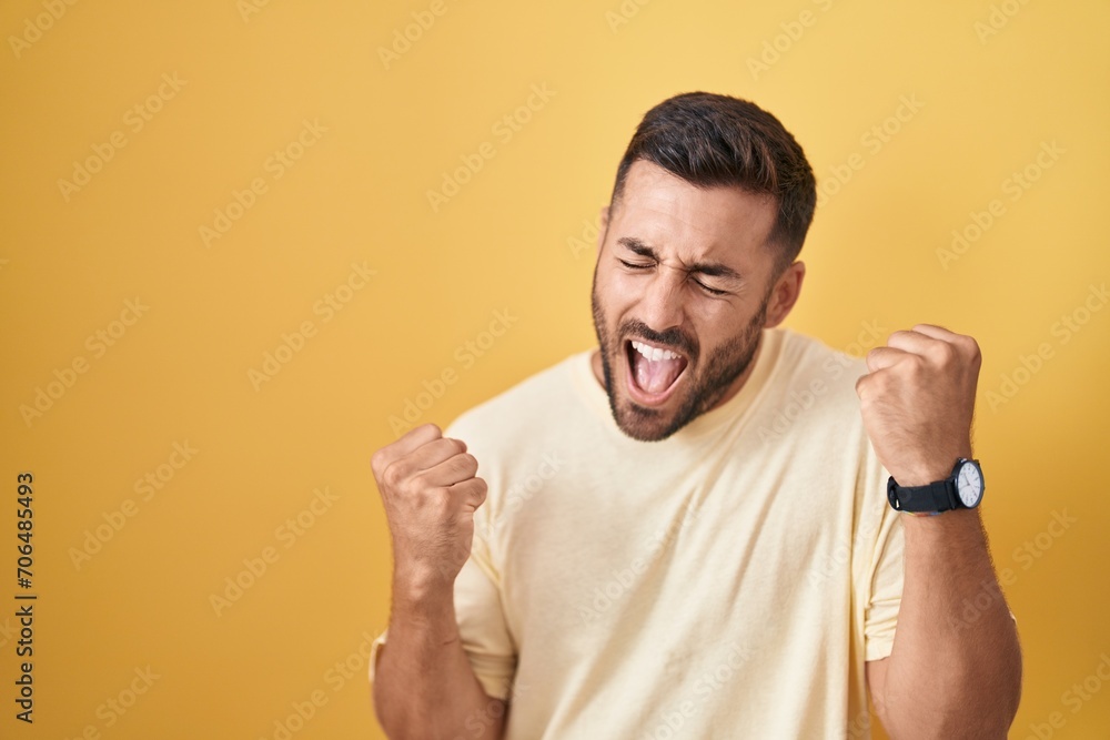 Handsome hispanic man standing over yellow background very happy and excited doing winner gesture with arms raised, smiling and screaming for success. celebration concept. Handsome hispanic man standing over yellow background very happy and excited doing winner gesture with arms raised, smiling and screaming for success. celebration concept.