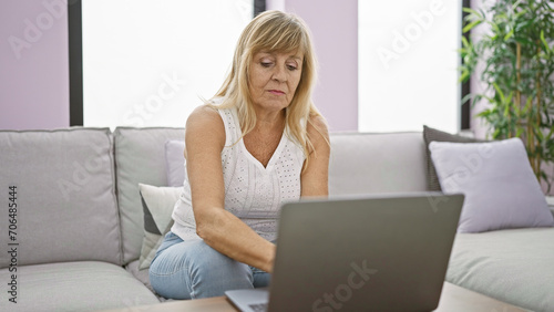 In her cozy home, a serious and focused middle age blonde woman is sitting on the sofa, totally absorbed, using her laptop in the living room's welcoming interior.