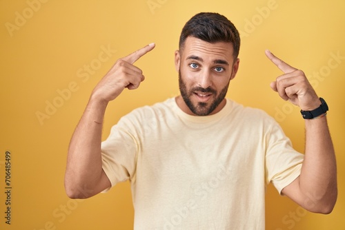 Handsome hispanic man standing over yellow background smiling pointing to head with both hands finger  great idea or thought  good memory