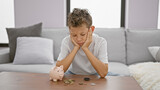 Adorable blond boy, sitting solemn and sad at home, sceptically studying coins and piggy bank, doubting the concept of saving