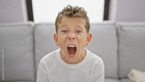 Adorable blond boy sitting on sofa at home, expressing loud frustration and anger photo
