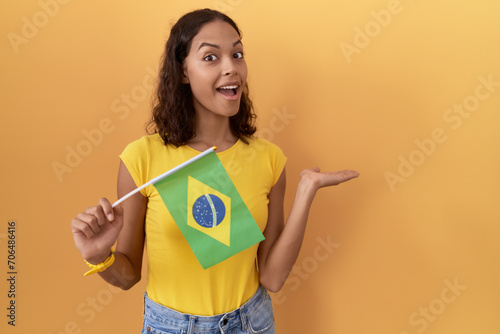 Young hispanic woman holding brazil flag pointing aside with hands open palms showing copy space, presenting advertisement smiling excited happy