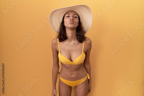 Young hispanic woman wearing bikini and summer hat looking at the camera blowing a kiss on air being lovely and sexy. love expression.