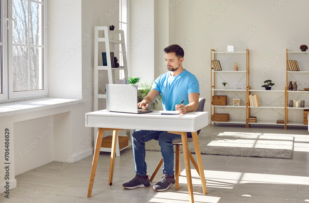 Portrait of young attractive business man wearing casual clothes working on a laptop at the office or at home, looking on a computer monitor screen and typing. Remote and freelance work concept.
