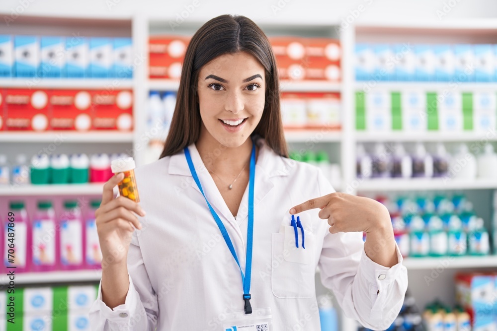 Hispanic woman working at pharmacy drugstore holding pills pointing finger to one self smiling happy and proud