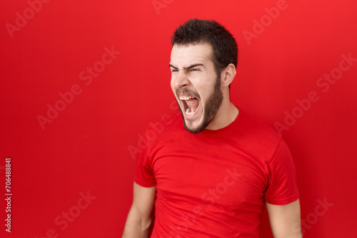 Young hispanic man wearing casual red t shirt angry and mad screaming frustrated and furious, shouting with anger. rage and aggressive concept.