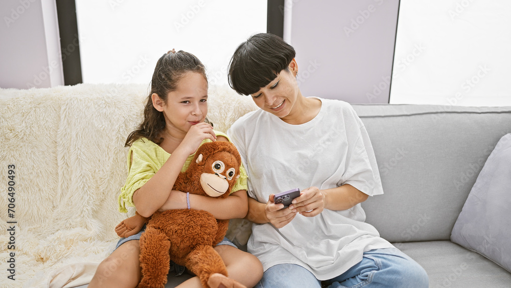 Casual fun at home, mother and daughter enjoy sitting on the sofa, hugging their lovely monkey doll while texting on their smartphone Casual fun at home, mother and daughter enjoy sitting on the sofa, hugging their lovely monkey doll while texting on their smartphone