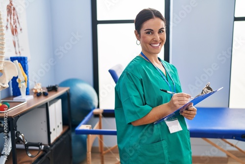 Young beautiful hispanic woman pysiotherapist smiling confident writing on document at rehab clinic