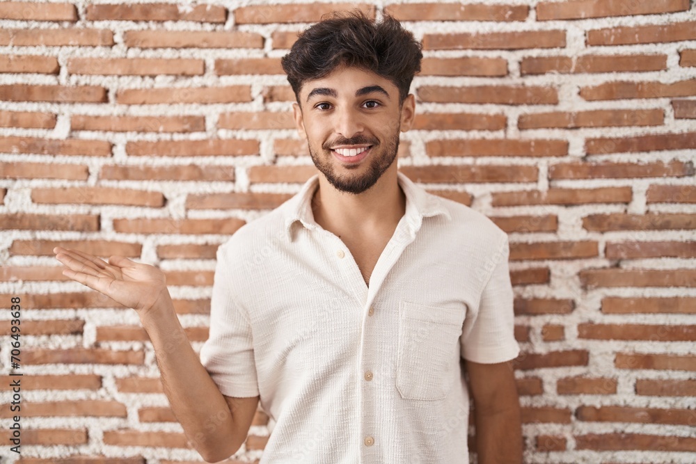 Arab man with beard standing over bricks wall background smiling cheerful presenting and pointing with palm of hand looking at the camera. Arab man with beard standing over bricks wall background smiling cheerful presenting and pointing with palm of hand looking at the camera.