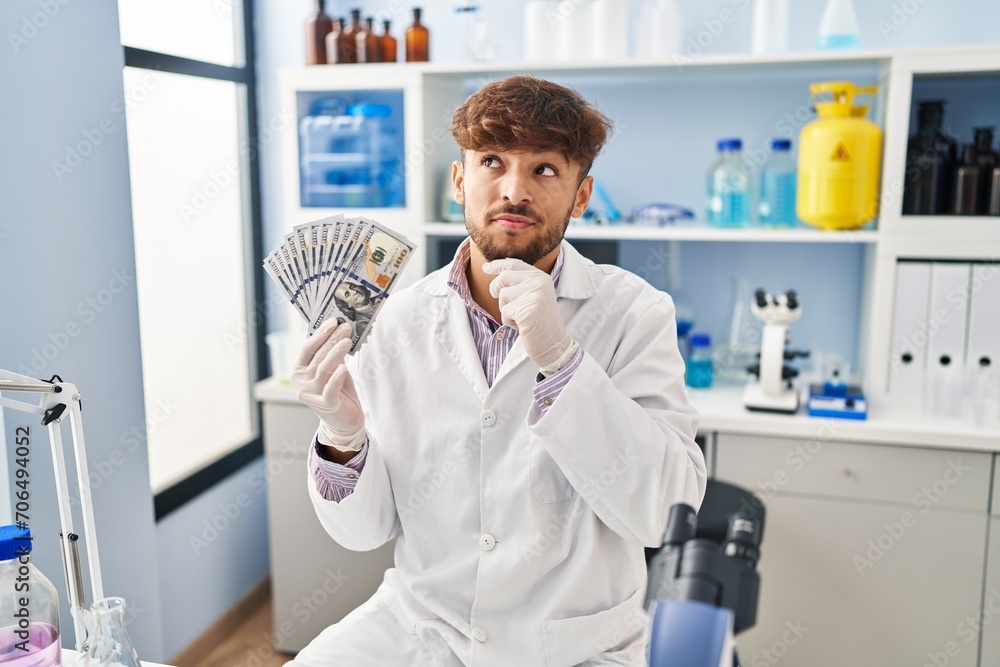 Arab man with beard working at scientist laboratory holding money serious face thinking about question with hand on chin, thoughtful about confusing idea