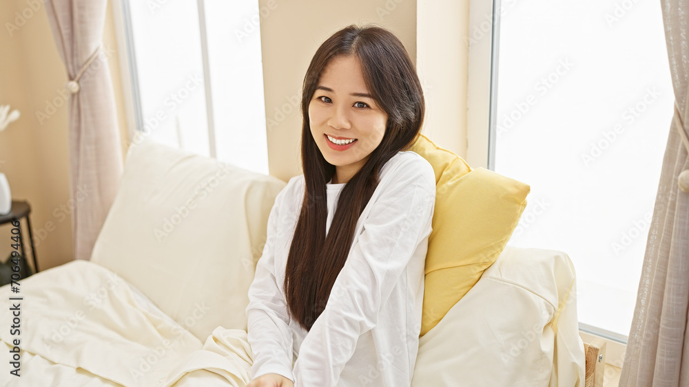 A cheerful young asian woman in a white shirt brightens a cozy, sunlit bedroom, embracing home comfort. A cheerful young asian woman in a white shirt brightens a cozy, sunlit bedroom, embracing home comfort.