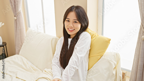 A cheerful young asian woman in a white shirt brightens a cozy  sunlit bedroom  embracing home comfort.