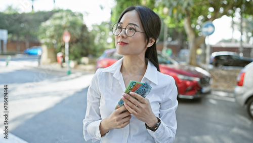An asian woman counts australian dollars on an urban street, reflecting city life and financial themes.