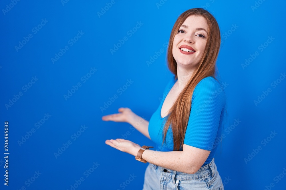 Redhead woman standing over blue background inviting to enter smiling natural with open hand Redhead woman standing over blue background inviting to enter smiling natural with open hand
