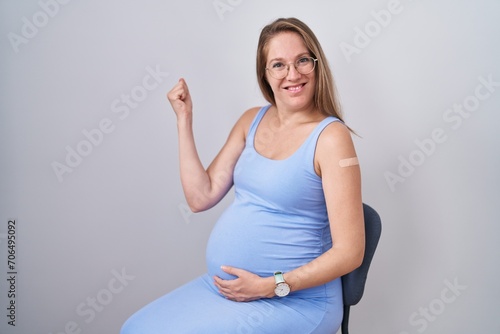 Young pregnant woman wearing band aid for vaccine injection screaming proud, celebrating victory and success very excited with raised arms