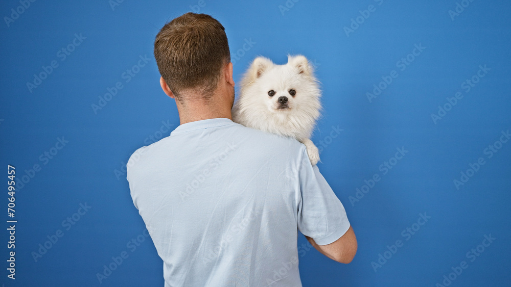 Handsome young caucasian man holding adorable pet dog, standing with back towards us, isolated against a neat outdoor blue wall background Handsome young caucasian man holding adorable pet dog, standing with back towards us, isolated against a neat outdoor blue wall background