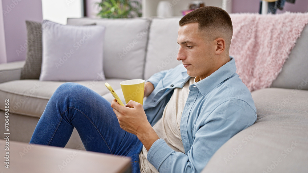 A young hispanic man relaxes at home on a sofa with a smartphone and coffee mug, showcasing casual, modern living.
