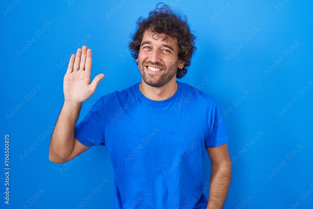 Naklejka premium Hispanic young man standing over blue background waiving saying hello happy and smiling, friendly welcome gesture