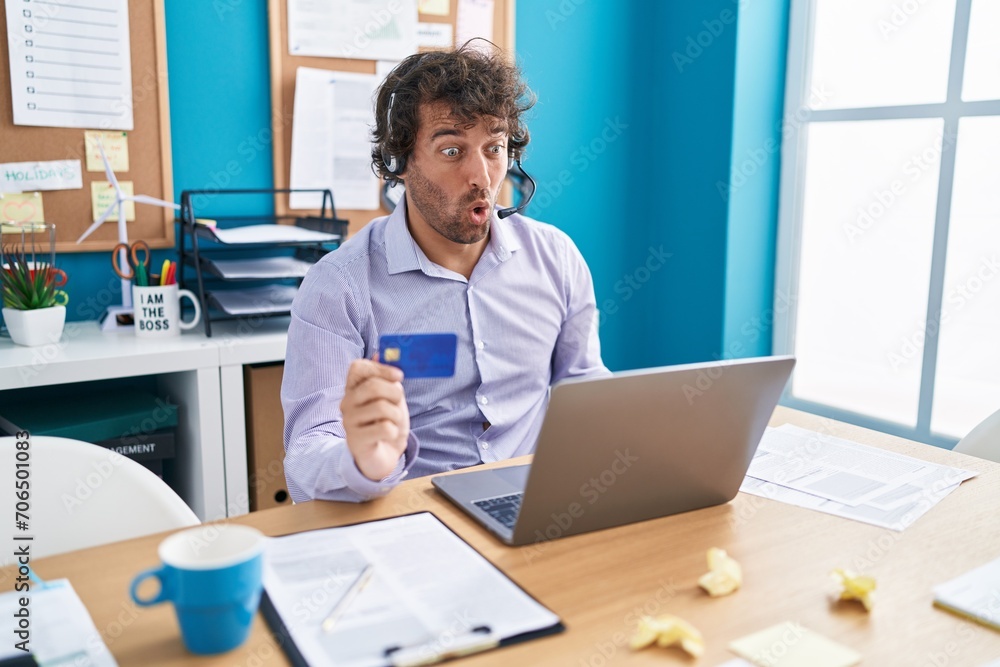 Hispanic young man working at the office doing online shopping scared and amazed with open mouth for surprise, disbelief face