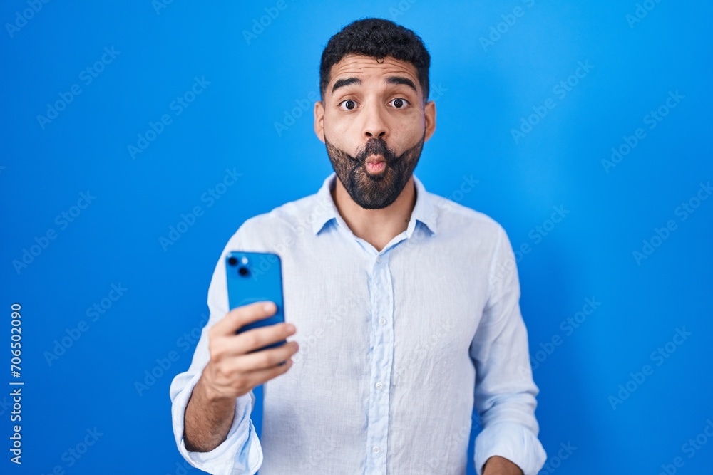 Hispanic man with beard using smartphone typing message making fish face with lips, crazy and comical gesture. funny expression. Hispanic man with beard using smartphone typing message making fish face with lips, crazy and comical gesture. funny expression.