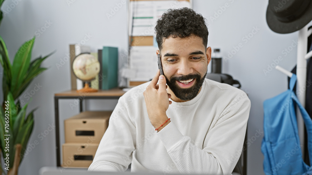 Fototapeta premium A smiling young hispanic man with a beard talking on a mobile phone in a modern office setting