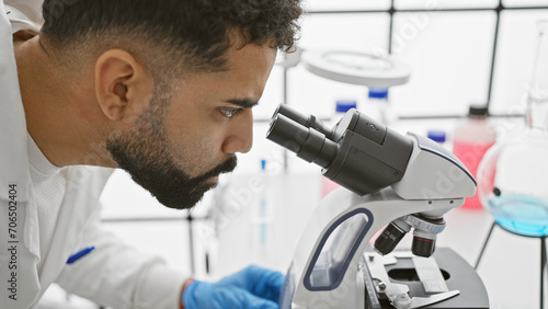 A focused young adult man with a beard wearing lab coat and gloves examines samples through a microscope in a modern laboratory.