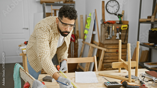 A young hispanic man wearing safety glasses measures wood in a well-organized carpentry workshop