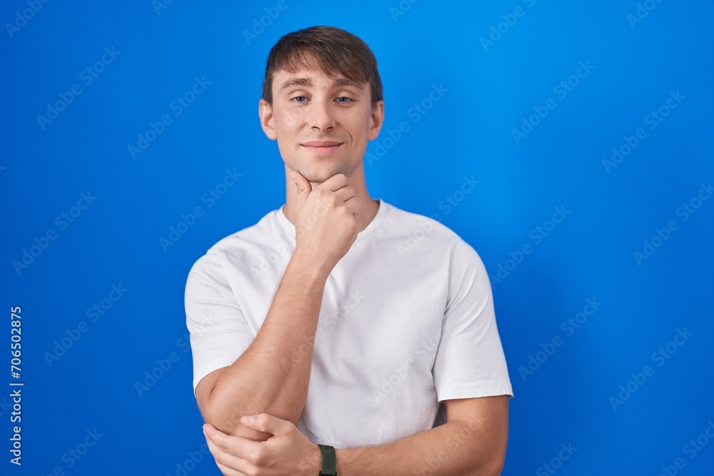 Caucasian blond man standing over blue background looking confident at the camera smiling with crossed arms and hand raised on chin. thinking positive. Caucasian blond man standing over blue background looking confident at the camera smiling with crossed arms and hand raised on chin. thinking positive.