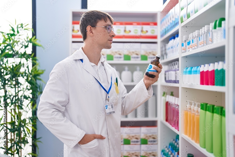 Young caucasian man pharmacist holding medication bottle of shelving at pharmacy