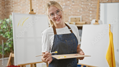 Young blonde woman artist smiling confident holding paintbrush and palette at art studio