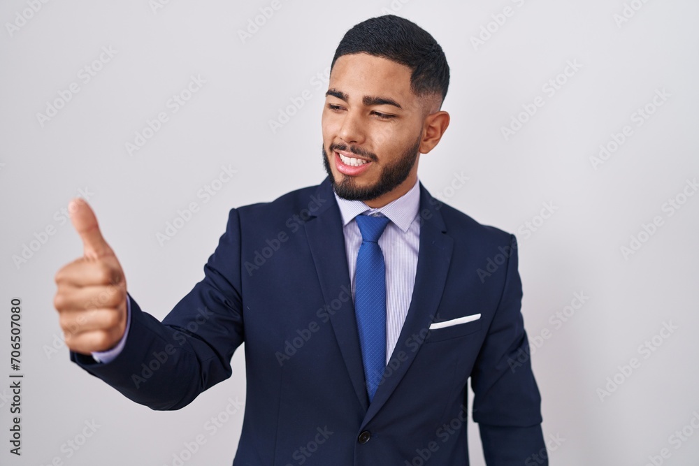 Young hispanic man wearing business suit and tie looking proud, smiling doing thumbs up gesture to the side Young hispanic man wearing business suit and tie looking proud, smiling doing thumbs up gesture to the side