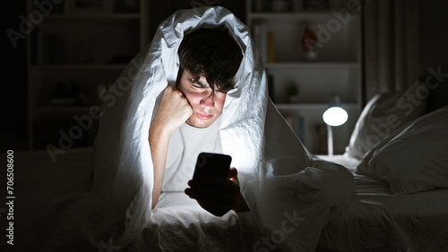 Young hispanic teenager seriously engrossed in texting on his smartphone, lying on a comfy bed under a snug blanket, in the dim light of his cozy bedroom