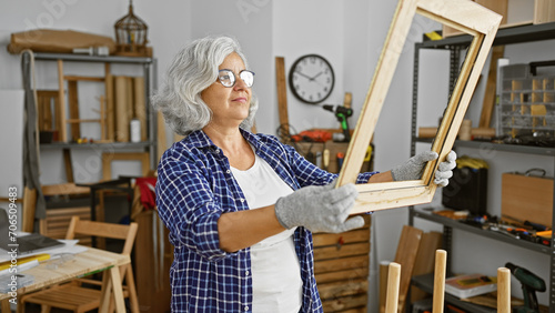 A mature woman examines a wooden frame in a well-organized carpentry workshop.