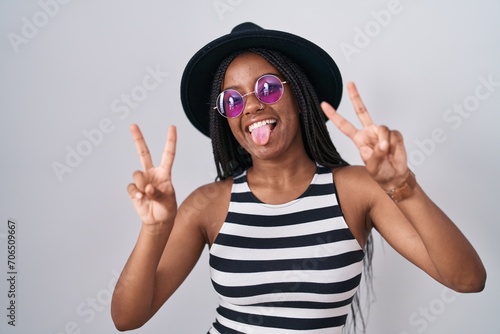 Young african american with braids wearing hat and sunglasses smiling with tongue out showing fingers of both hands doing victory sign. number two.