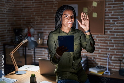 Young african american with braids working at the office at night waiving saying hello happy and smiling, friendly welcome gesture photo
