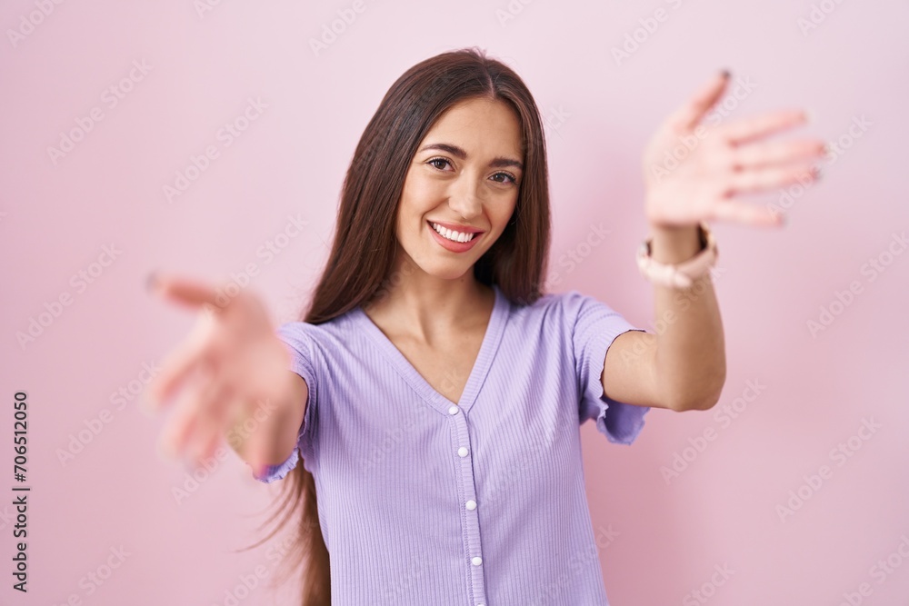 Young hispanic woman with long hair standing over pink background looking at the camera smiling with open arms for hug. cheerful expression embracing happiness. Young hispanic woman with long hair standing over pink background looking at the camera smiling with open arms for hug. cheerful expression embracing happiness.